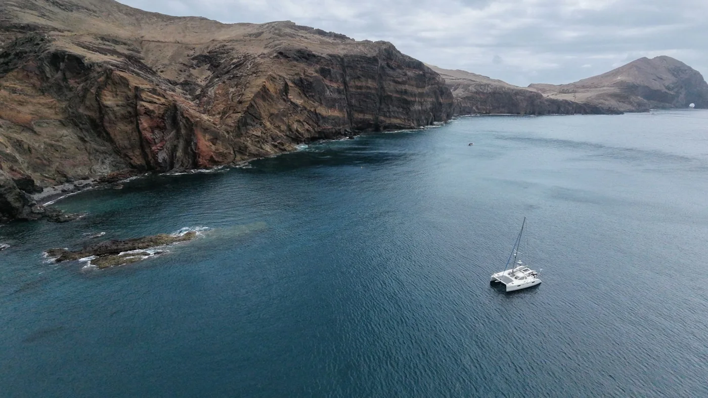 Little Cloud, our Catana 431 sailing catamaran, anchored alone under high layered volcanic cliffs